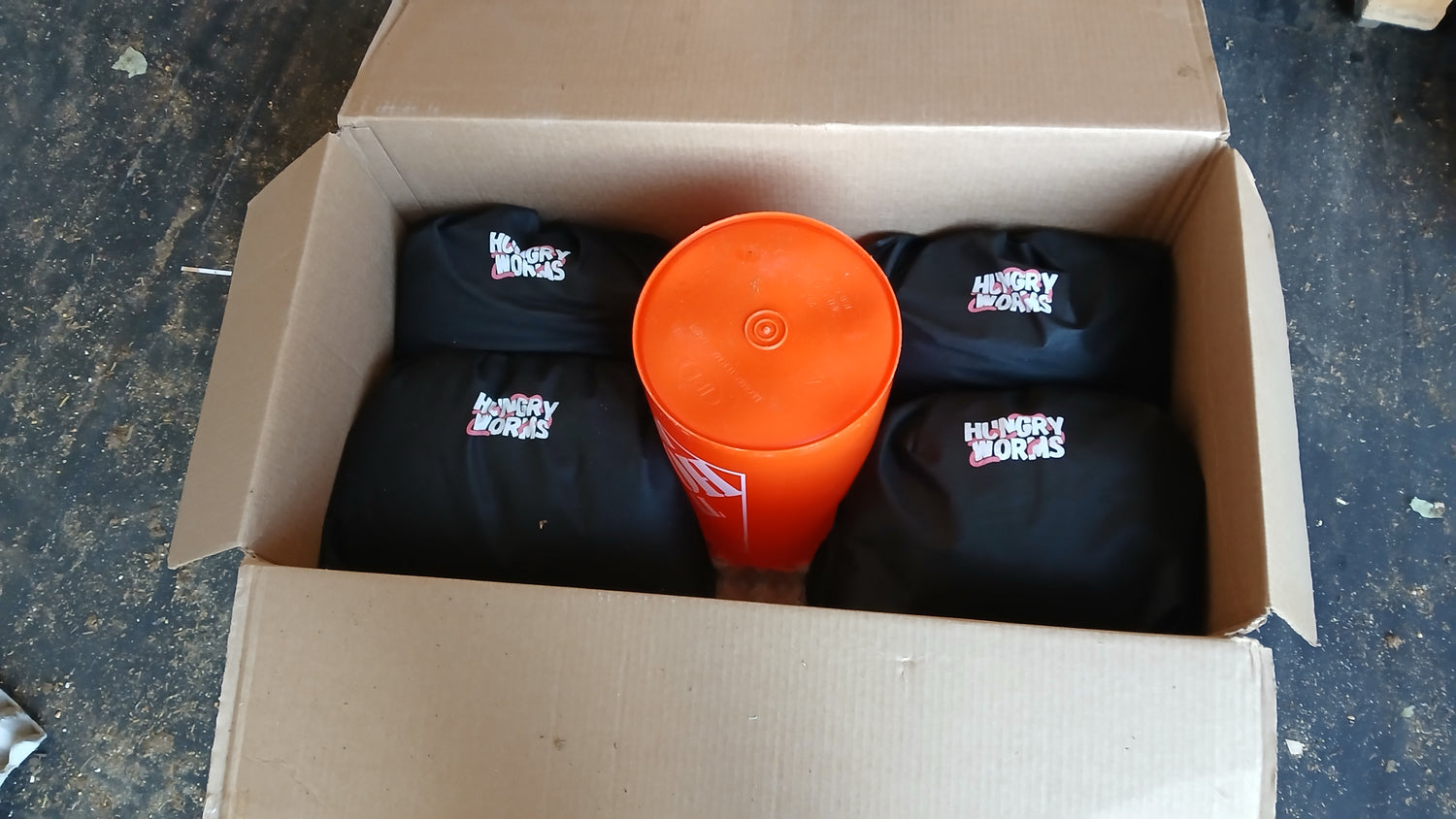 Box containing black bags with visible Hungry Worms branding and an orange bucket inside to protect the package containing worms in shipment